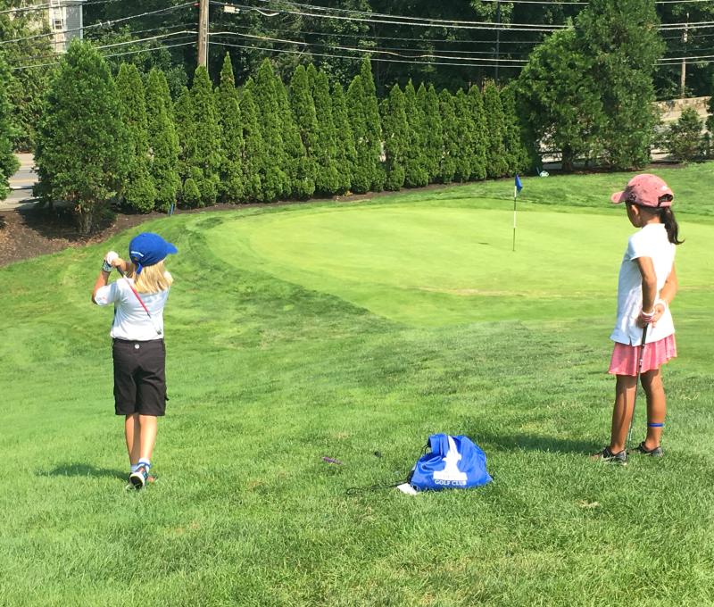 One junior golfer swings while the other junior golfer watches